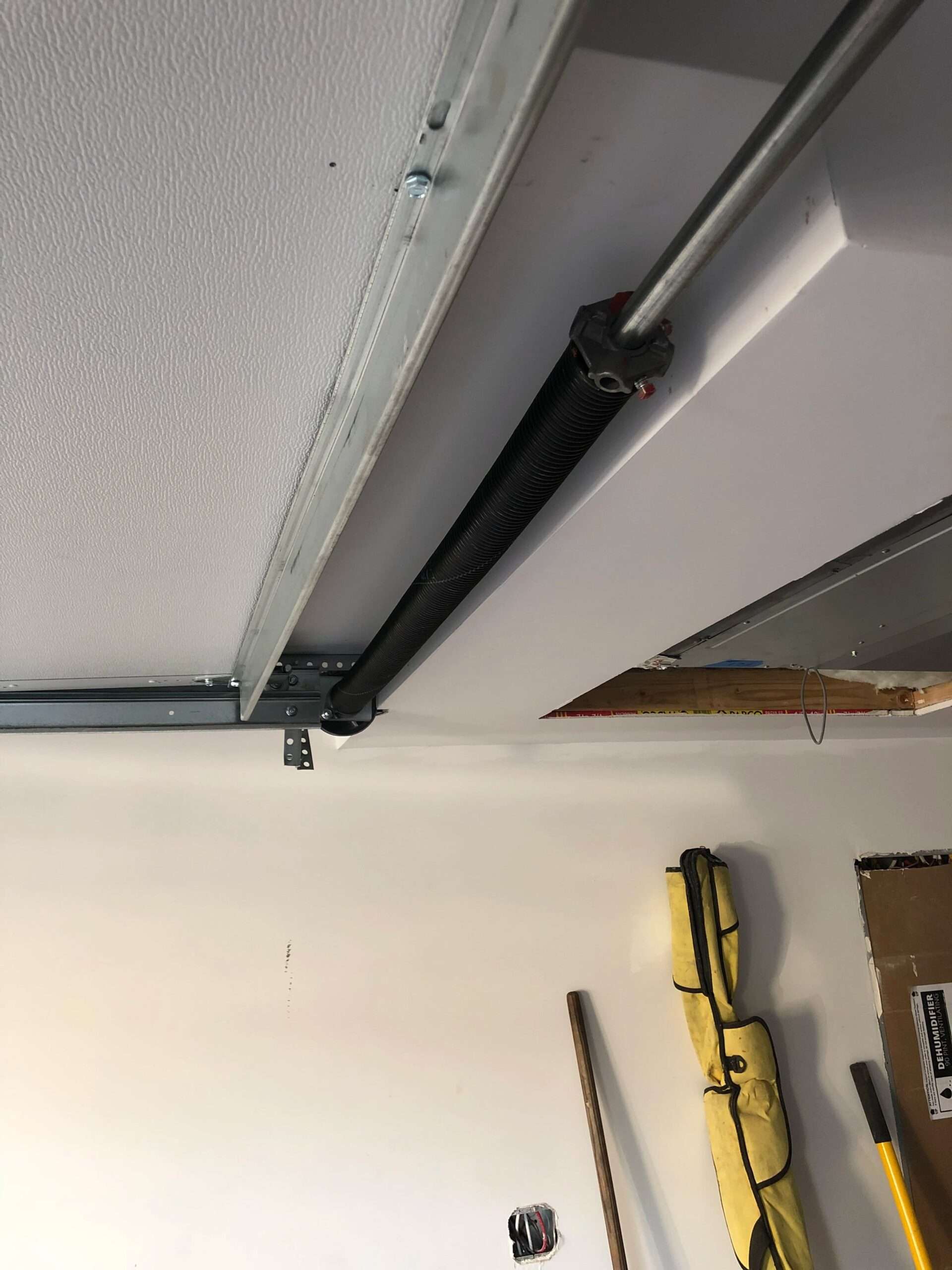 REVERSE-TORSION Close-up of a garage door torsion spring and track mounted on a ceiling, with tools and gloves visible on a nearby wall.