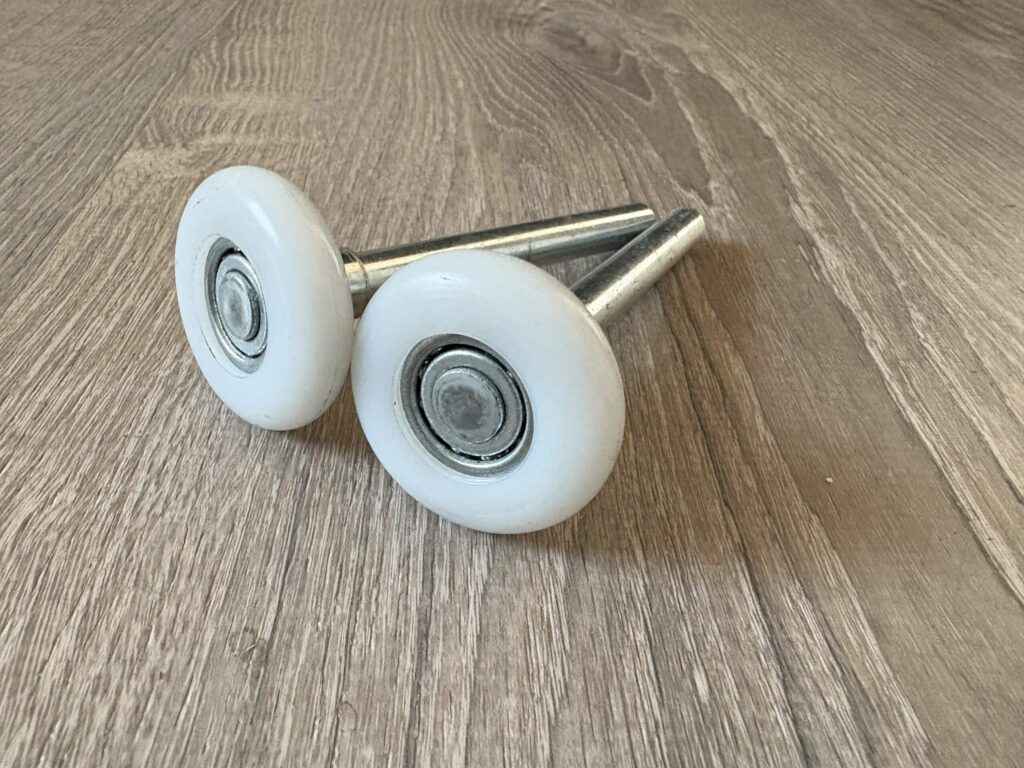 Two white garage door rollers with metal axles on a light wooden surface,
